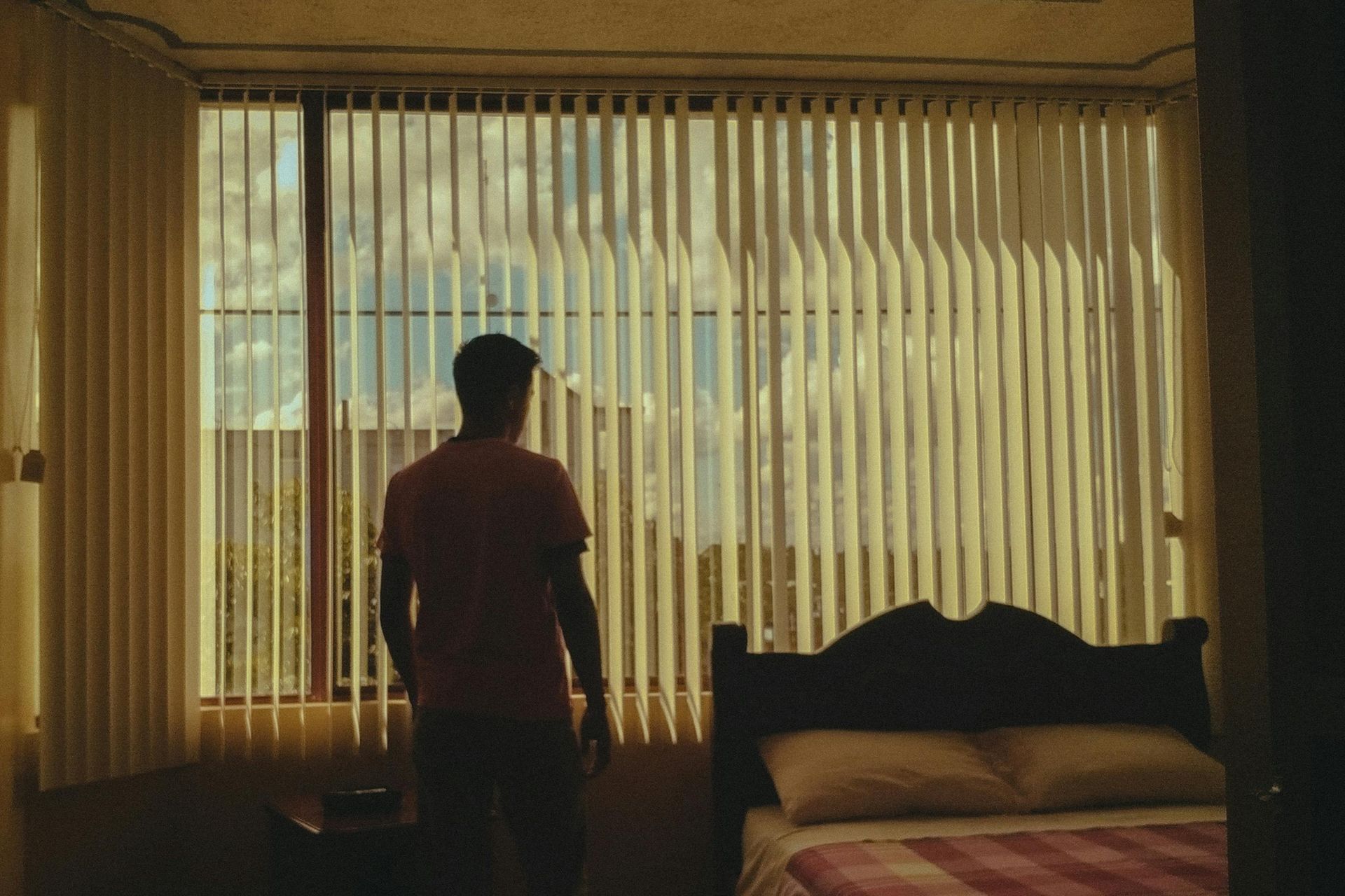 Man looking out a window with vertical blinds, sunlight streaming in. Bedroom setting, bed visible.