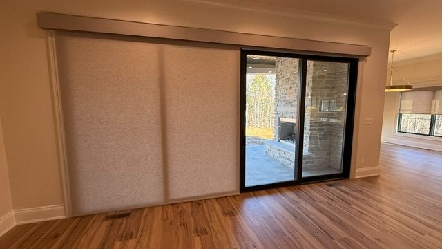 Empty living room with wood floors, beige walls, and sliding glass doors with roller shades opening to a patio.