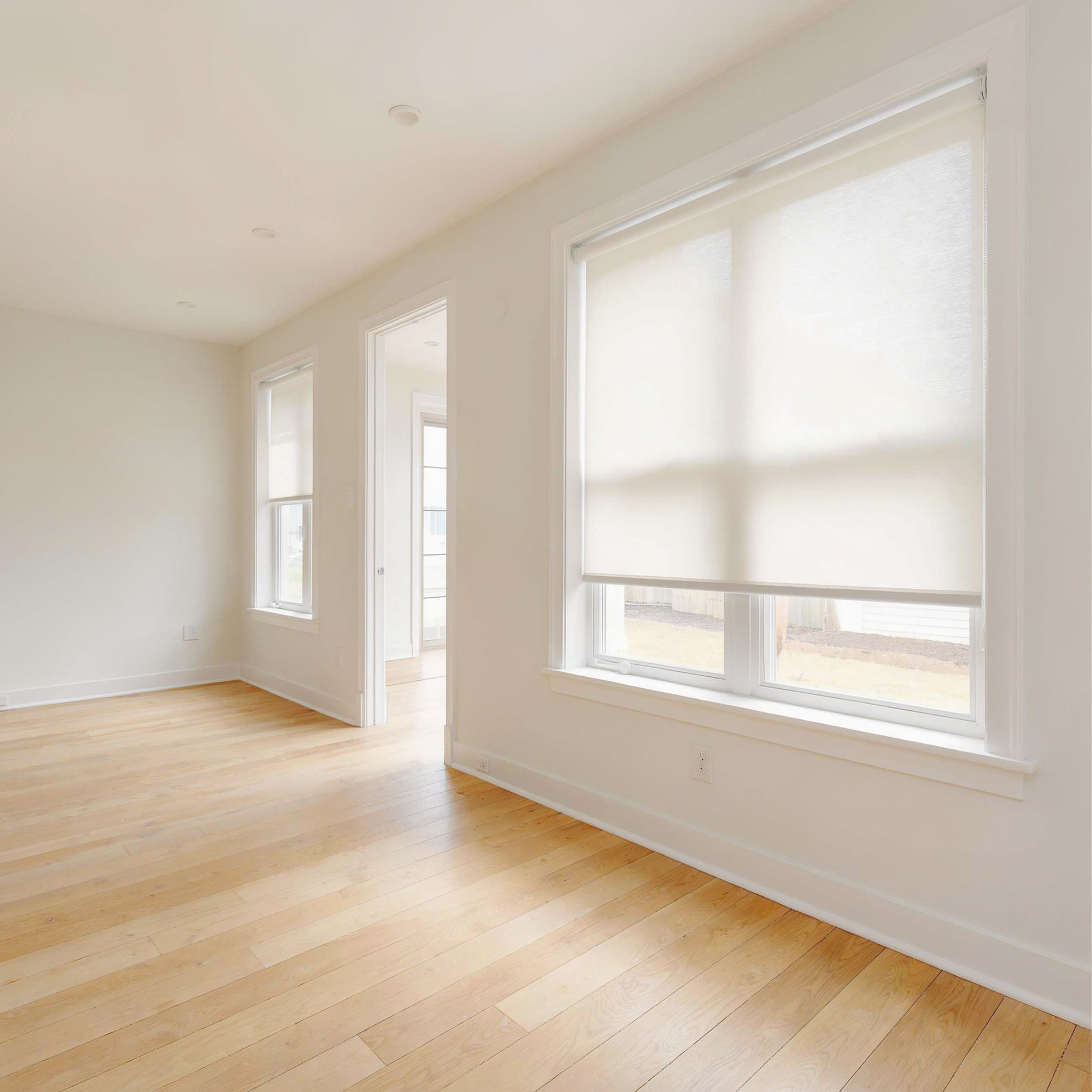 Bright empty room with light wood floors, white walls, and three tall windows with motorized shades.