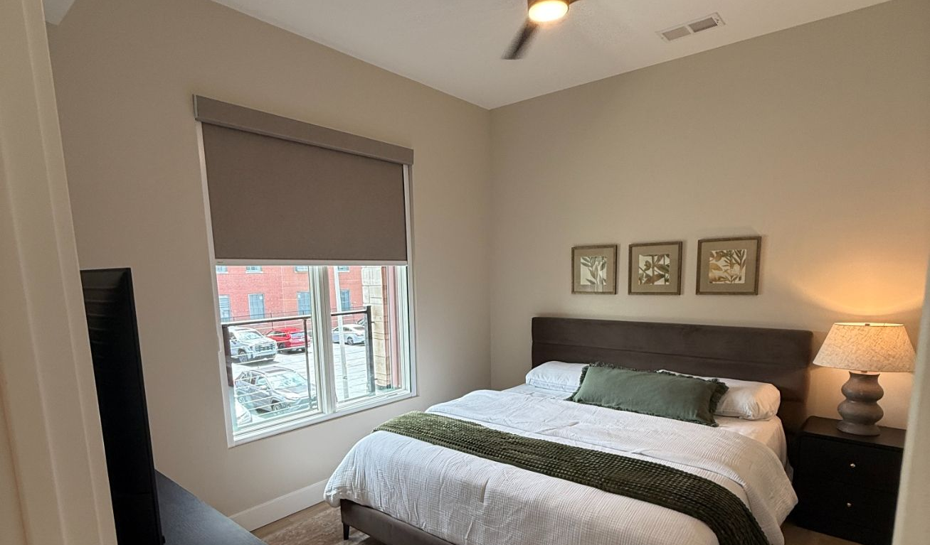 Cozy bedroom with a bed, gray walls, bedside lamp, and a window with motorized roller shades.