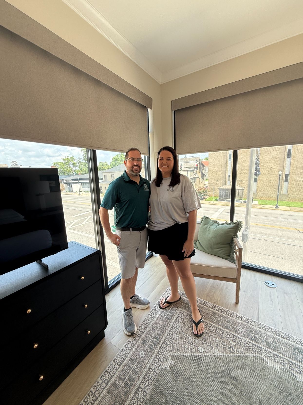 Two people standing together in a bright living room beside a TV and large window.
