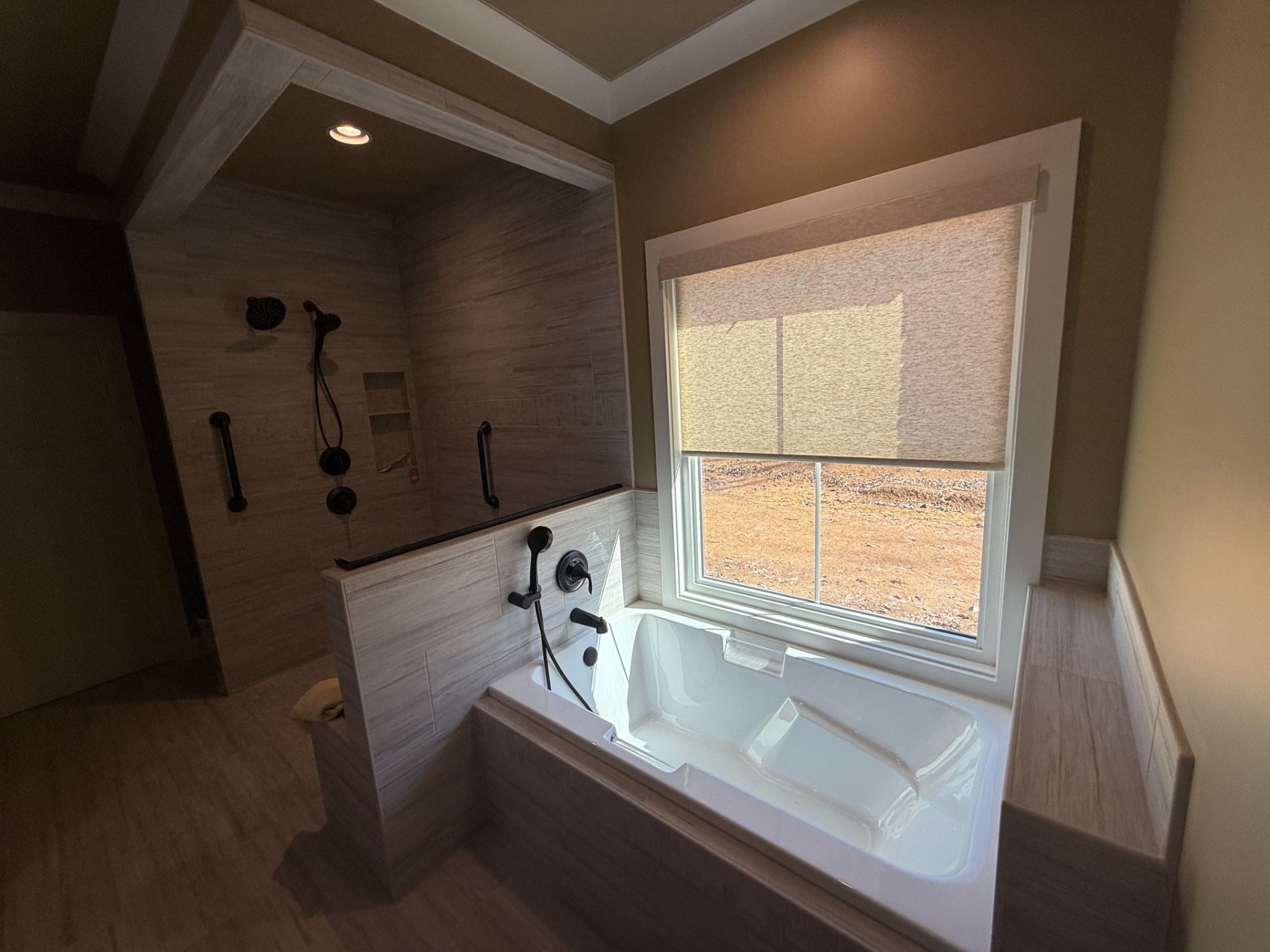Modern bathroom with a built-in tub, large window, and tiled shower wall in warm neutral tones.
