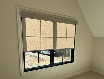 Beige motorized window shade partially lowered over a black-framed window in a light-colored room.