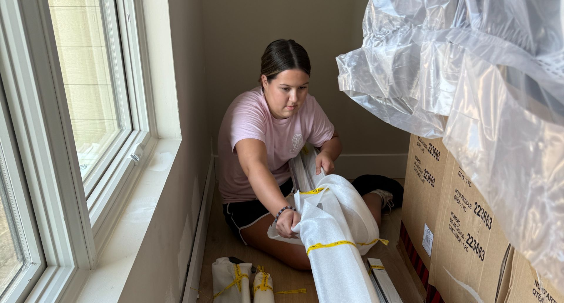 Simmonds family member packing a large rolled item in a windowed room, surrounded by cardboard boxes and plastic covering.
