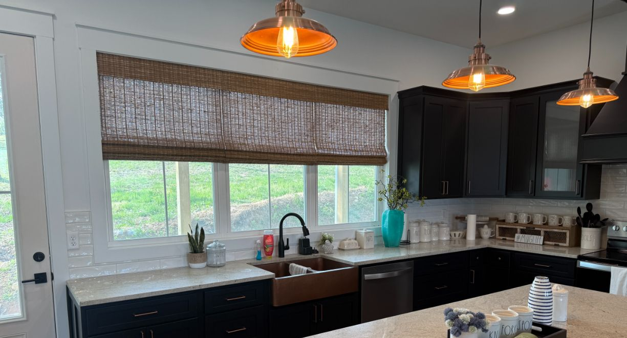 Modern kitchen with black cabinets, white countertops, woven wood shades, and a large window over the sink.