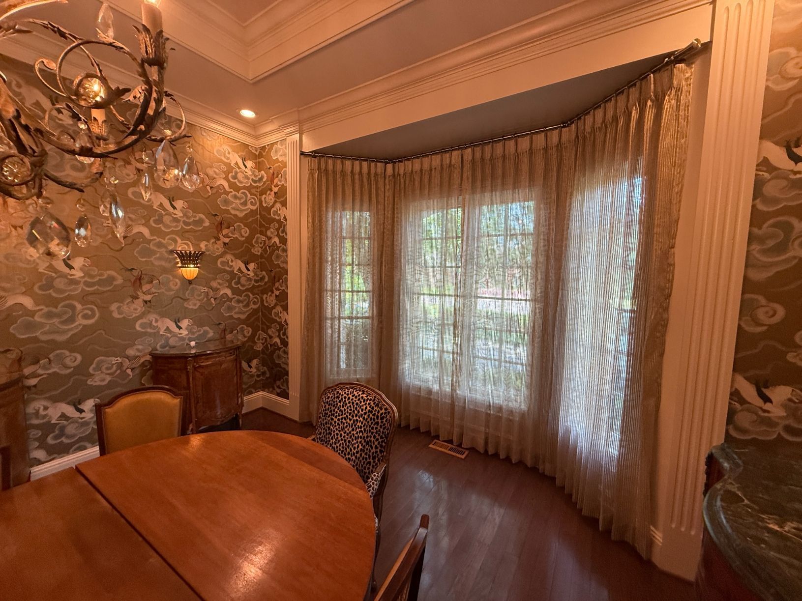 Elegant dining room with floral wallpaper, wood table, patterned chairs, and sunlight through sheer drapery.