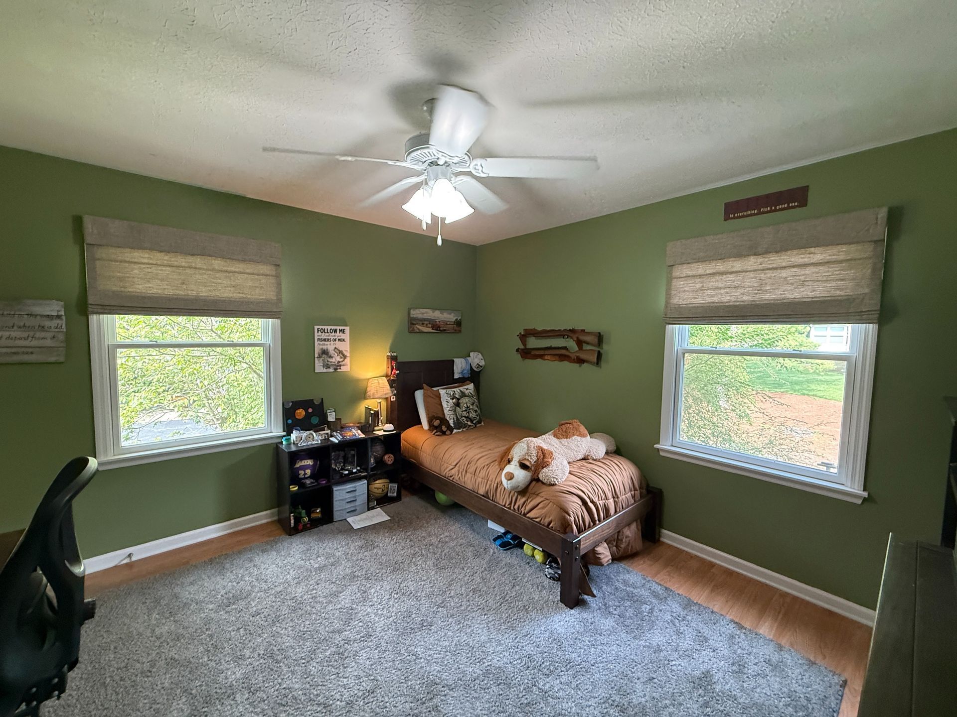 Green bedroom with twin beds, two windows with roman shades, ceiling fan, and gray carpeted floor.
