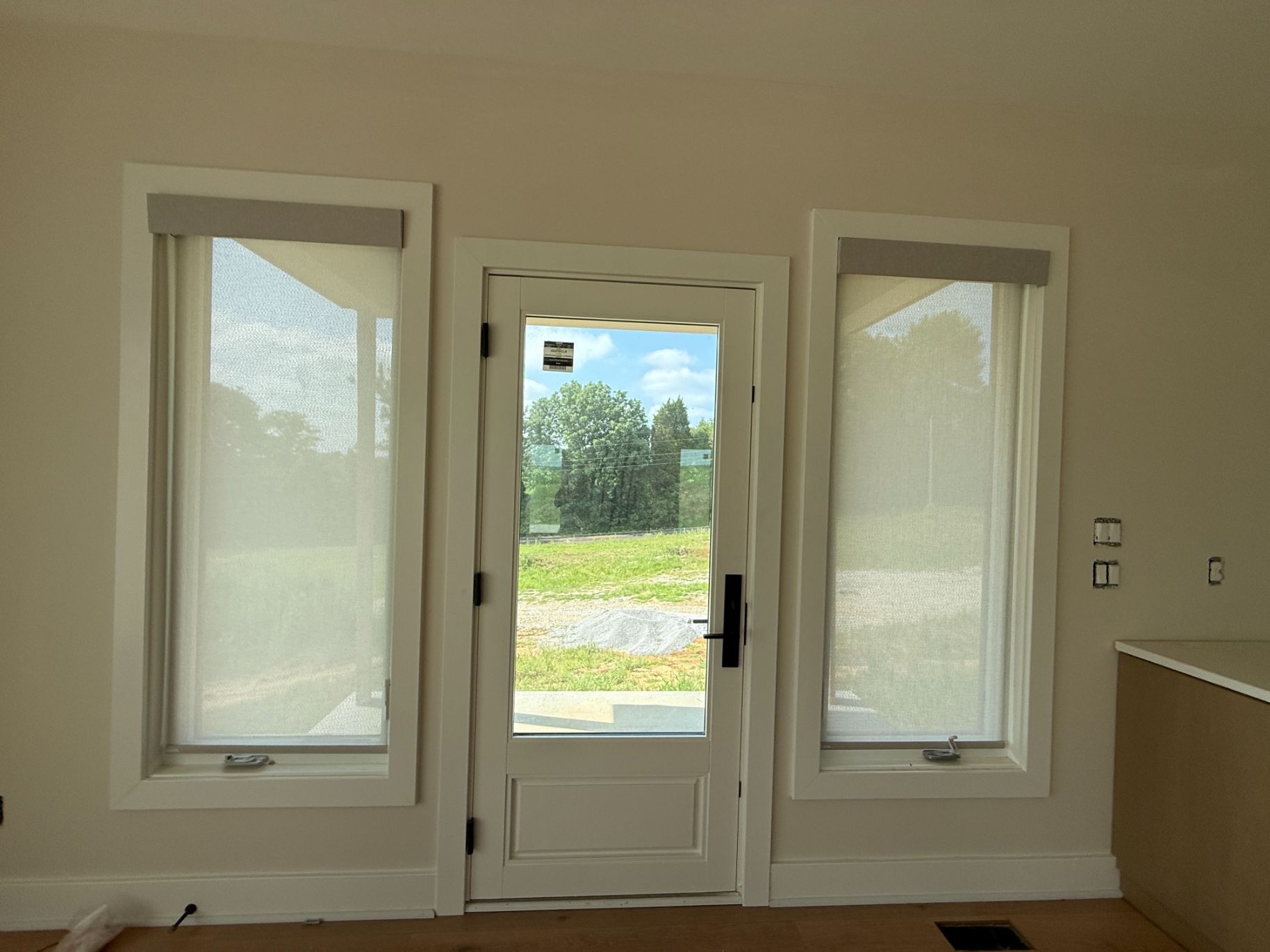 Bright interior with front door flanked by two windows with solar shades, looking out to a grassy yard and trees.