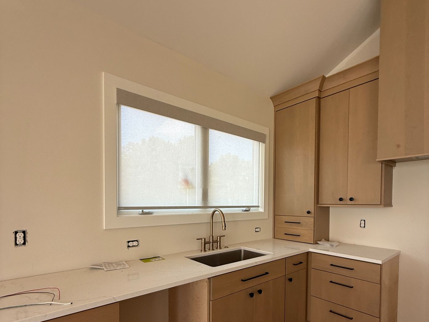 Empty kitchen with light wood cabinets, white countertops, and a large window with roller shades above the sink.