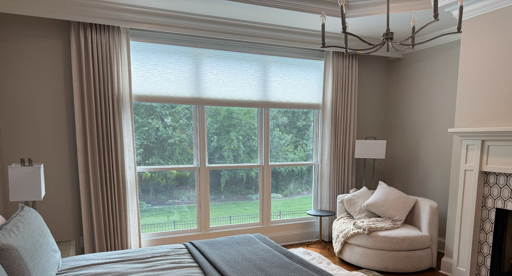 Bright living room with large window, beige drapery, honeycomb shades, fireplace, and green yard view