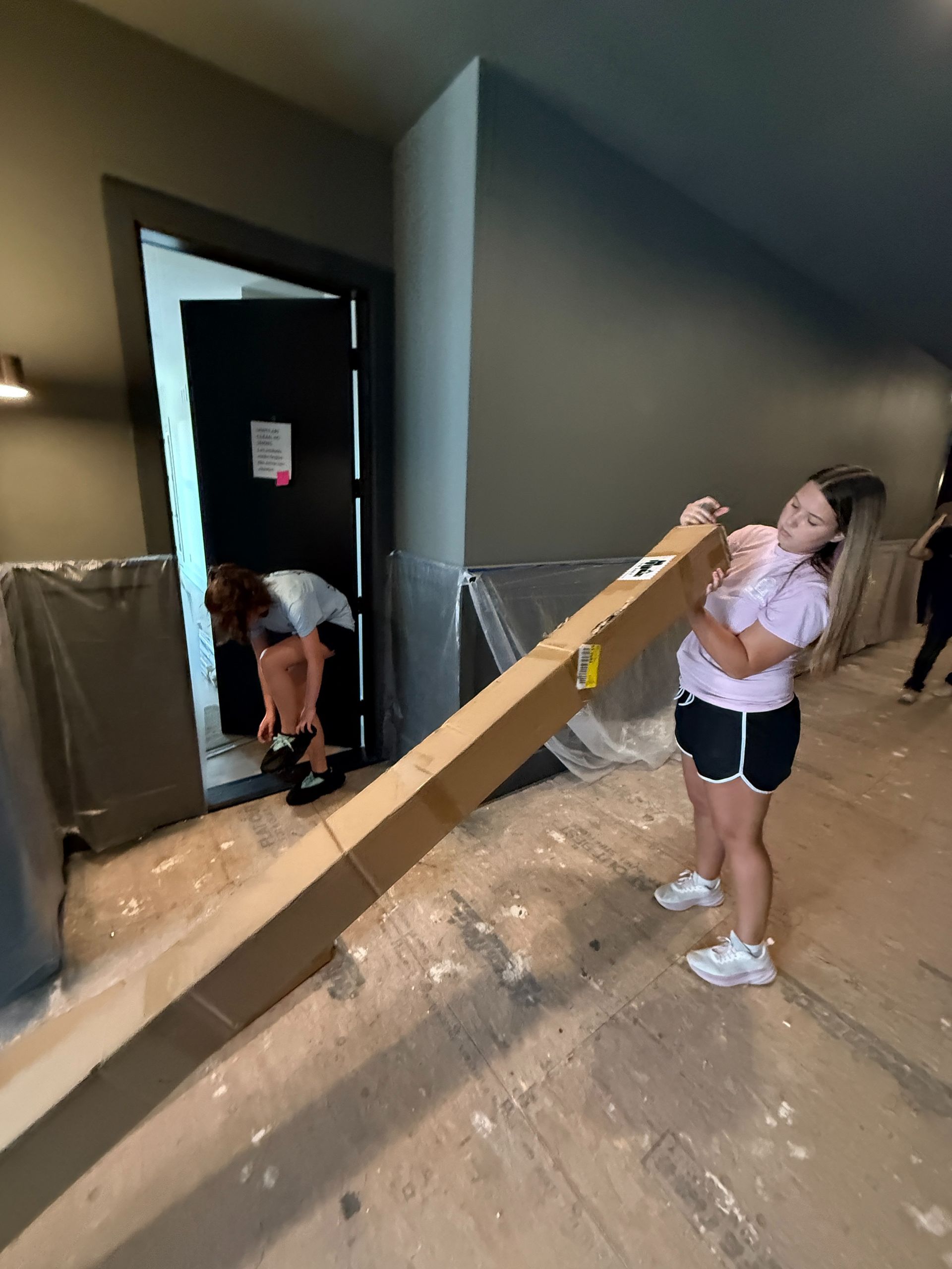 Simmonds family members moving a long cardboard boxes through a doorway in a messy indoor space
