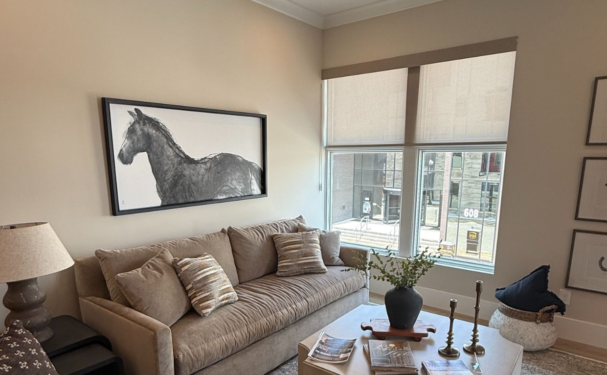 Cozy living room with beige sofa, patterned chair, coffee table, wall art, and large window with roller shades.