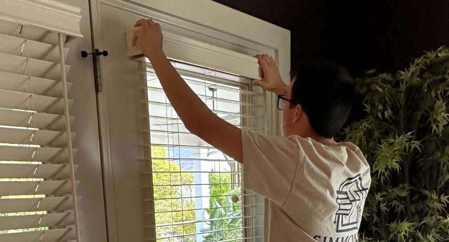 Simmonds family member adjusting white blinds on a window in a dim room .