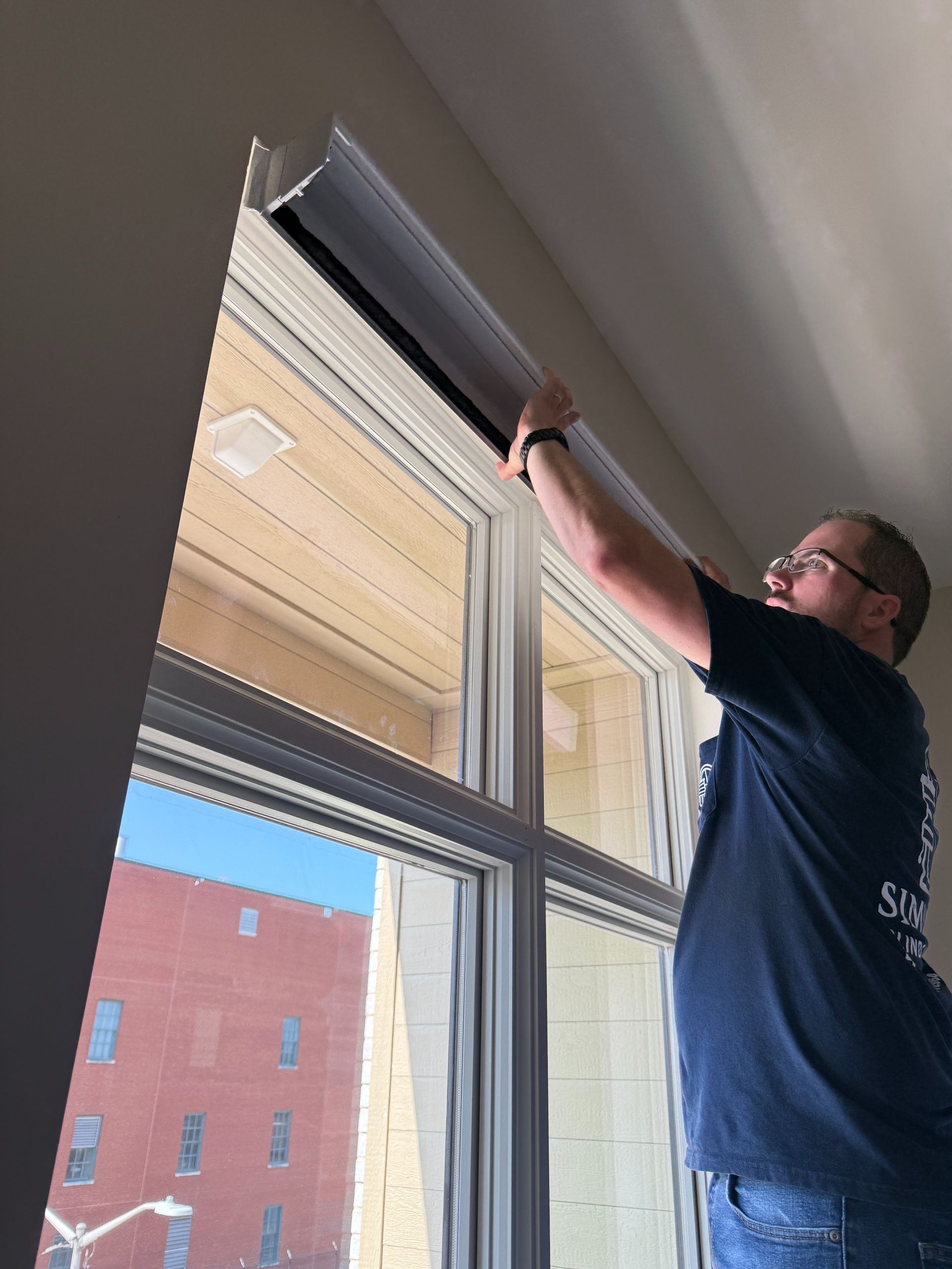 Sean Simmonds is adjusting a black window blind indoors beside a large window