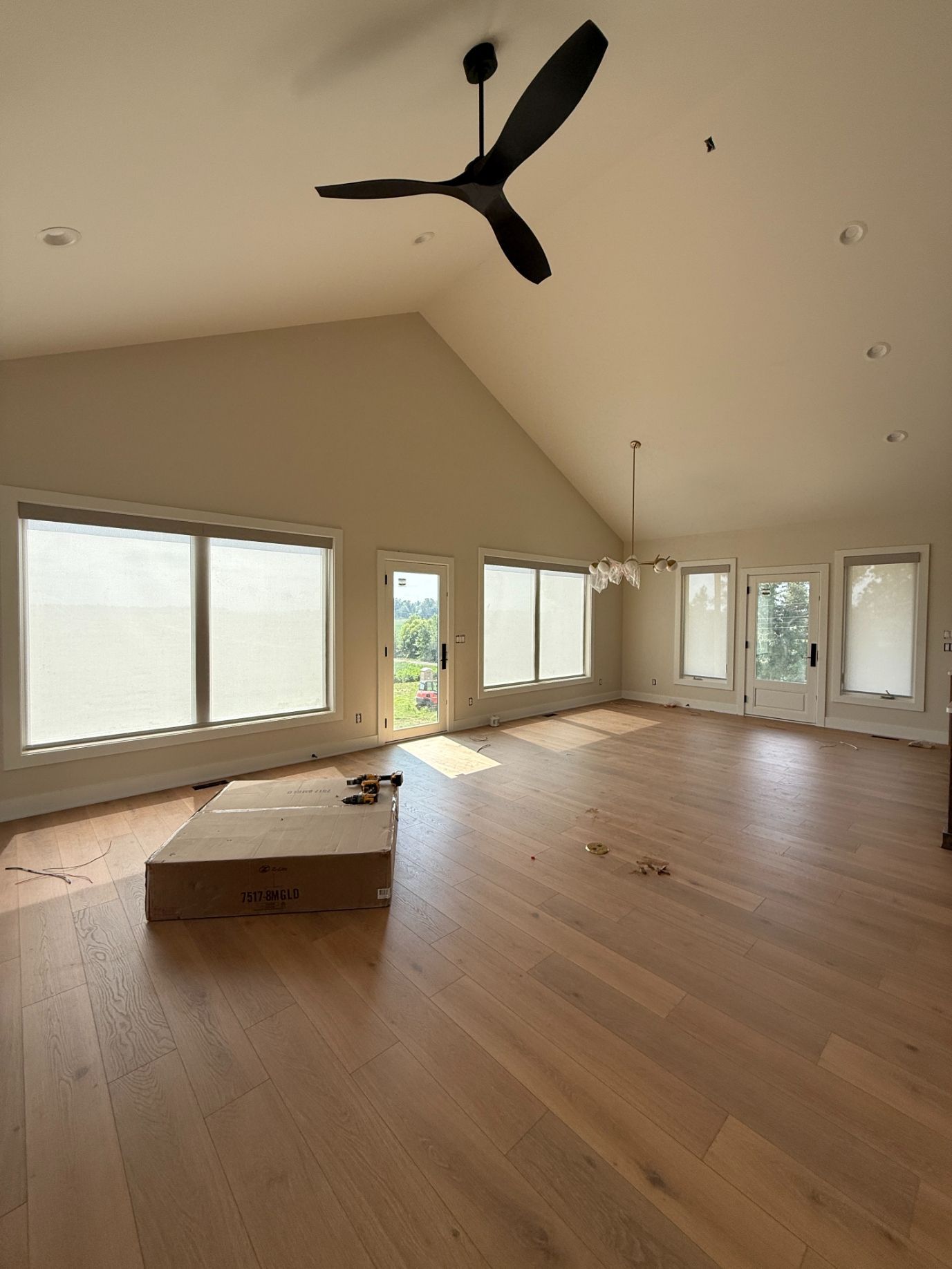 Bright empty living room with wood floors, large windows with roller shades, and a ceiling fan
