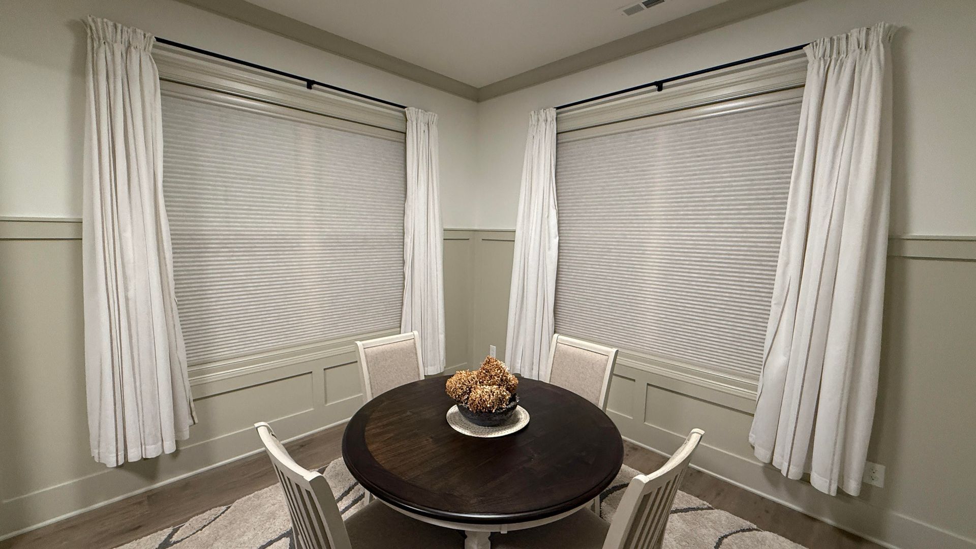 Dining area with round table, four chairs, and two windows with white curtains.