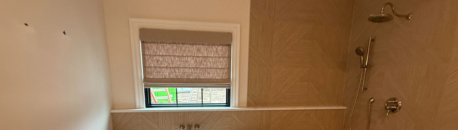 A bathroom with a window and a shower with a gold finish. The walls are tiled with a similar color.