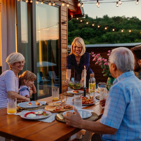 Friends dining outdoors on a patio at sunset, sharing food and drinks under string lights.