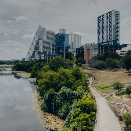 Riverfront city skyline with modern high-rise buildings, a winding path, and trees along the water