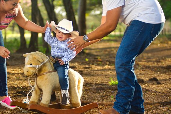 Child in a cowboy hat riding a toy horse with adults helping outdoors in a park