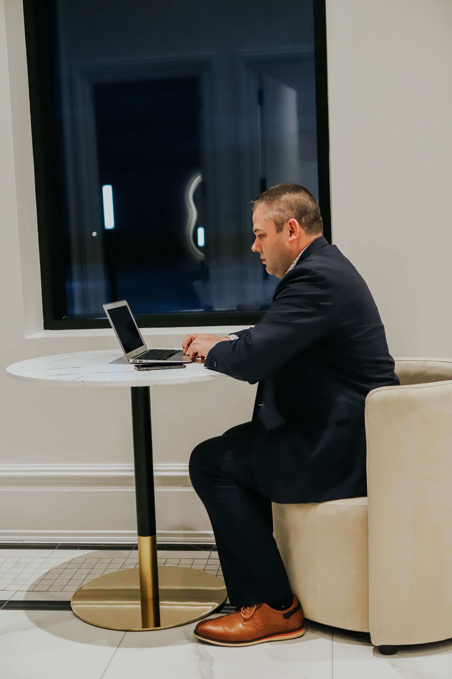 Man in a dark suit working on a laptop at a small round table in a modern indoor lounge