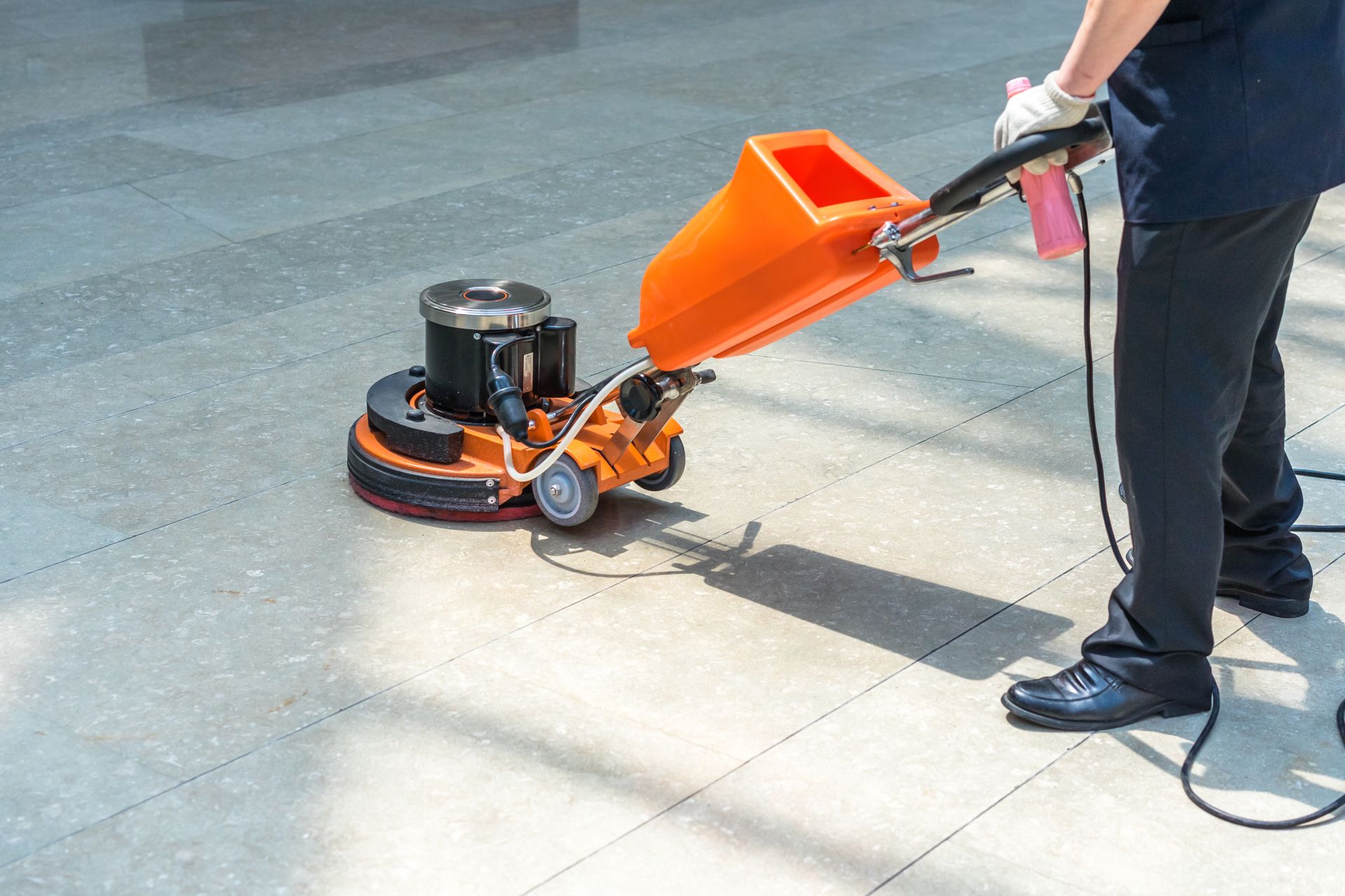 Worker using an orange floor grinder on a concrete surface outdoors.