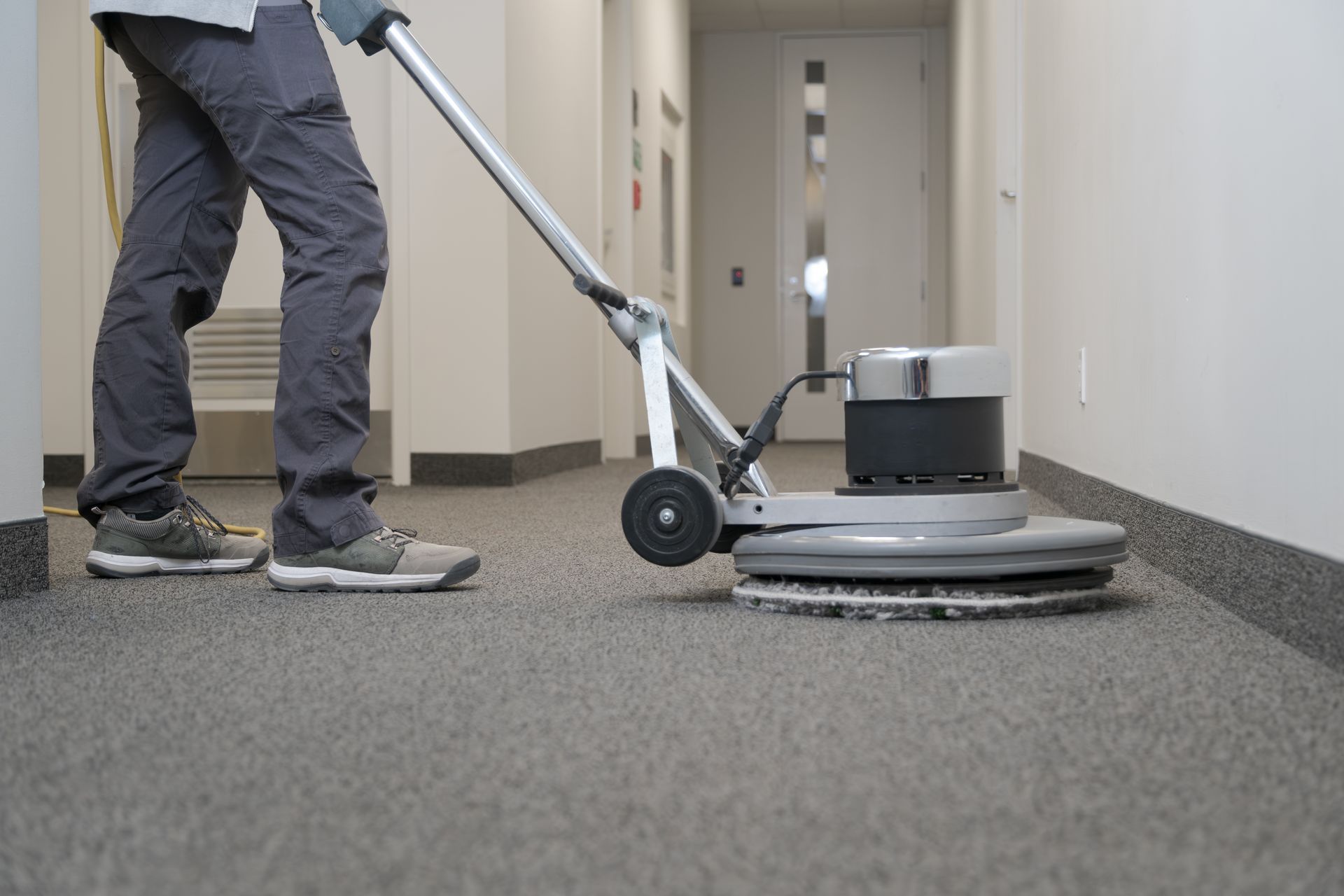 Person operating a floor buffer in a hallway with gray carpet and white walls Person operating a floor buffer in a hallway with gray carpet and white walls