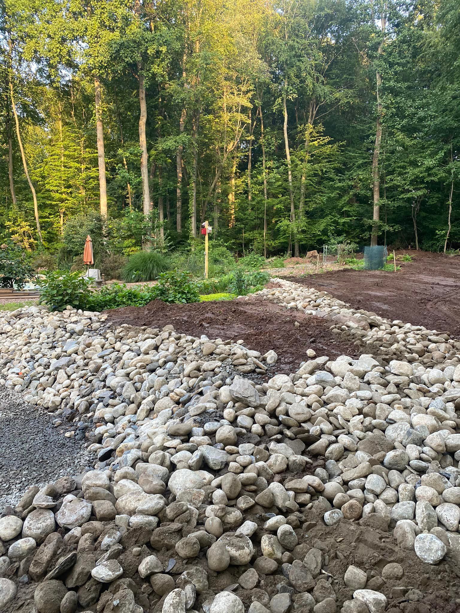 Rocky landscaping with freshly tilled soil, a garden with plants, and a forest backdrop.