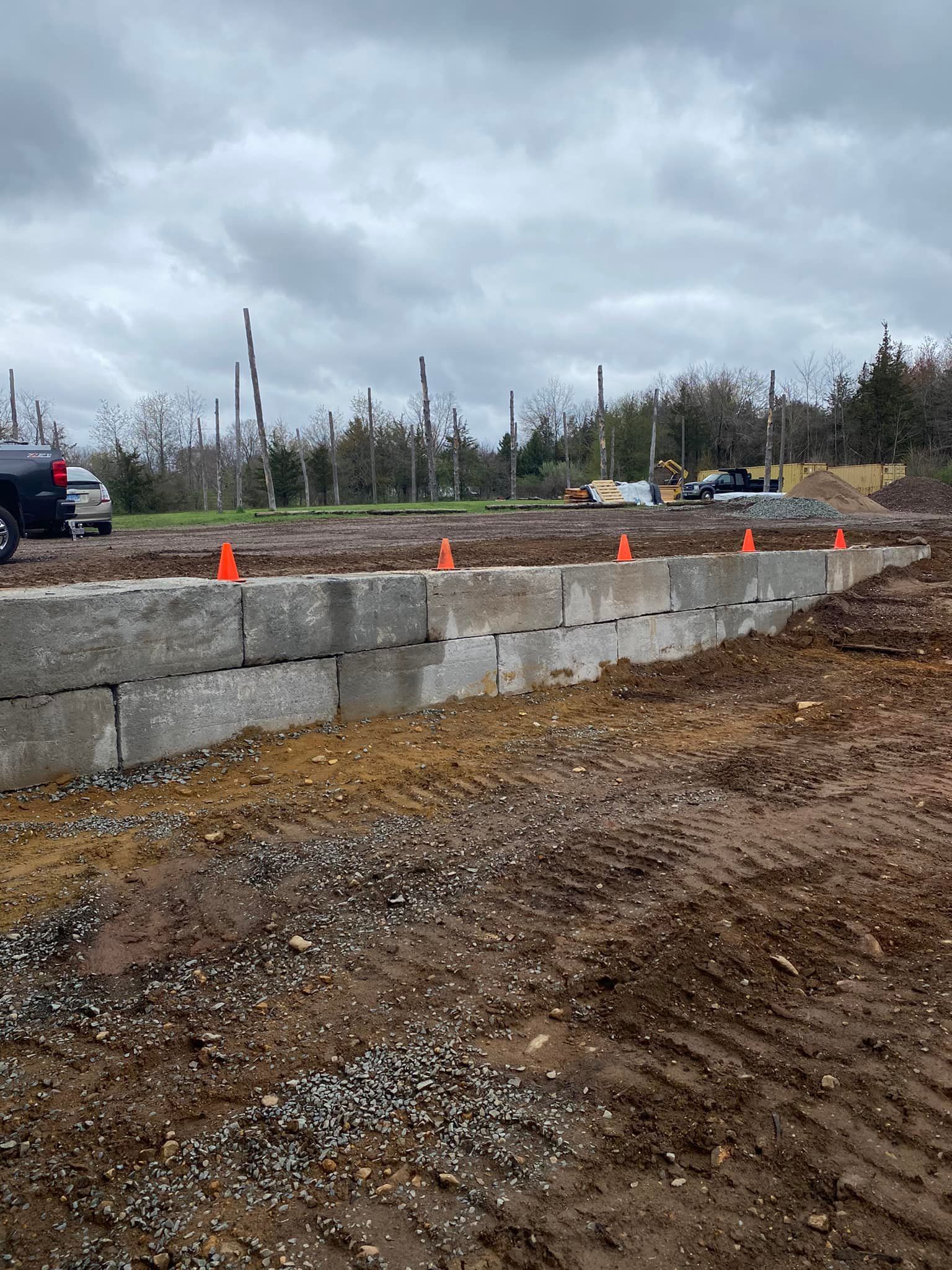 Concrete block retaining wall at a construction site, topped with orange cones. Cloudy sky.