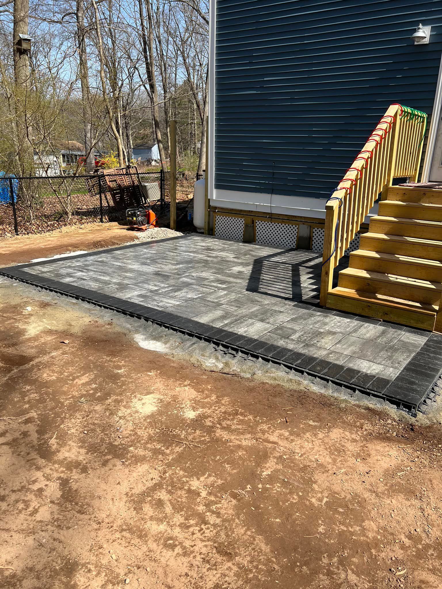 Newly constructed stone patio with stairs leading to a blue-sided house. Black edging and tan dirt surround.