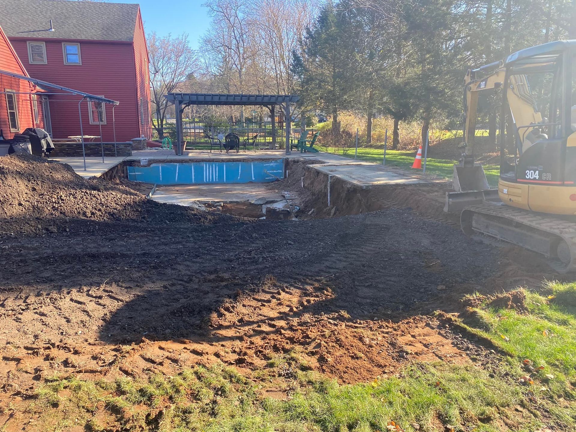 Excavation for a pool in a backyard, with a partially installed blue pool. Red house and pergola in the background.