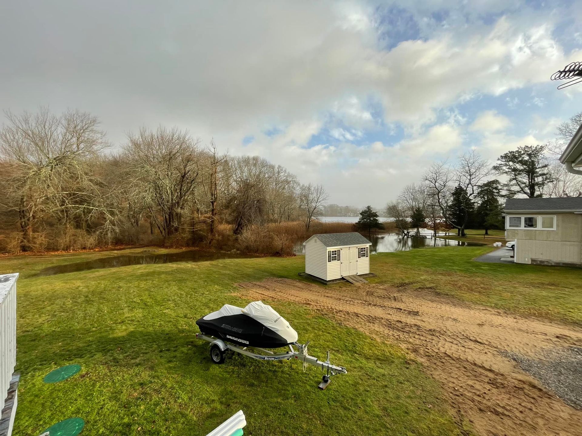 Grassy yard with a jet ski on a trailer, small shed, and trees leading to a body of water under a cloudy sky.