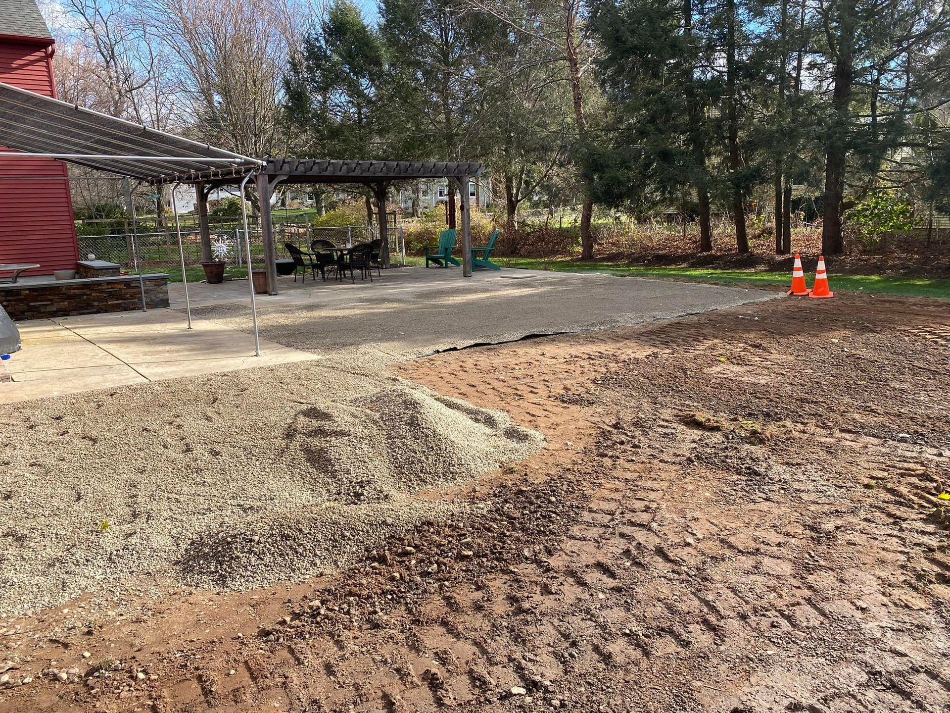 Gravel patio under construction near a wooden pergola and red building; dirt and tire tracks in foreground.