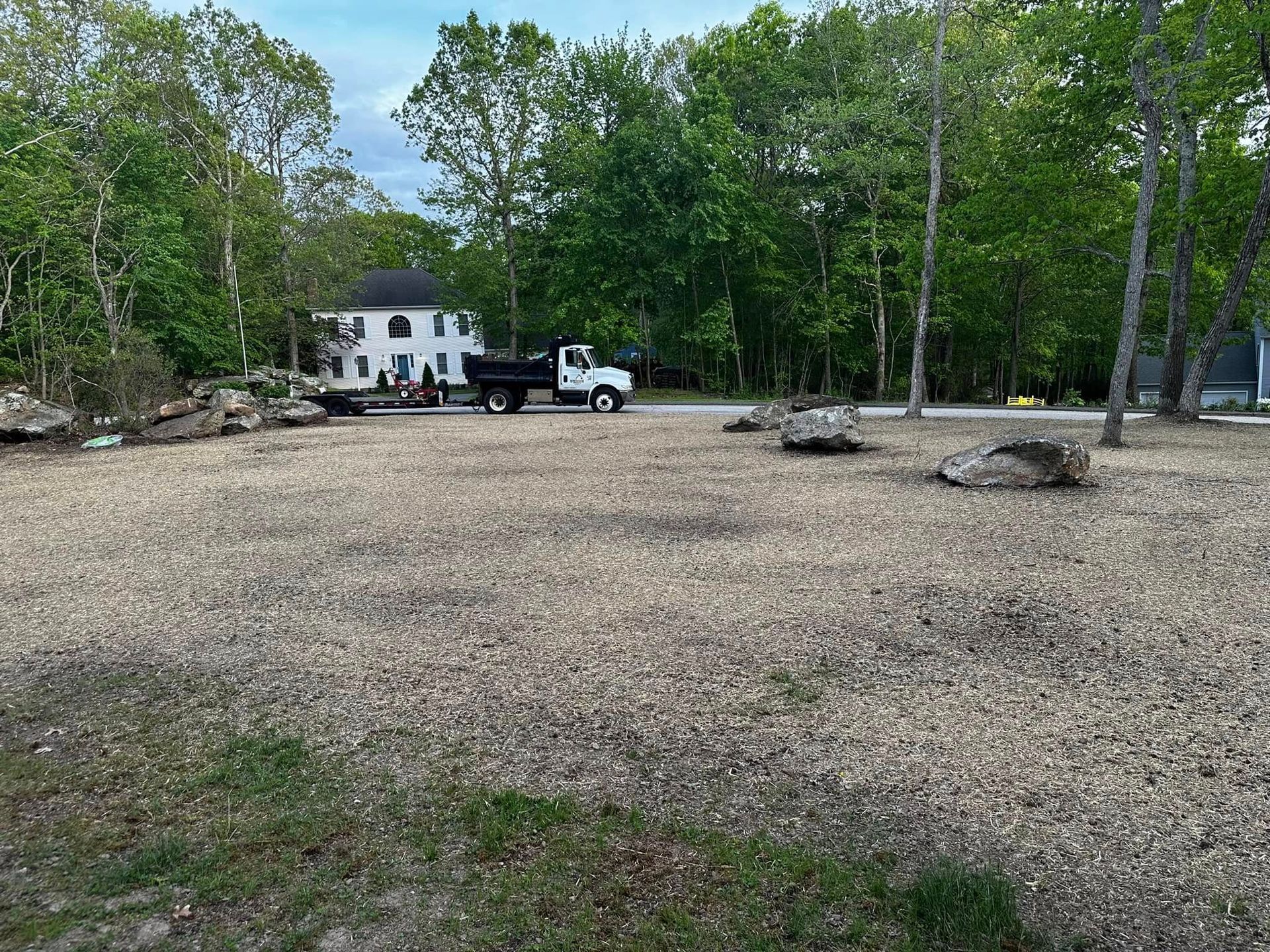 Gravel lot with large rocks, a white truck, and a two-story white house in a wooded area.