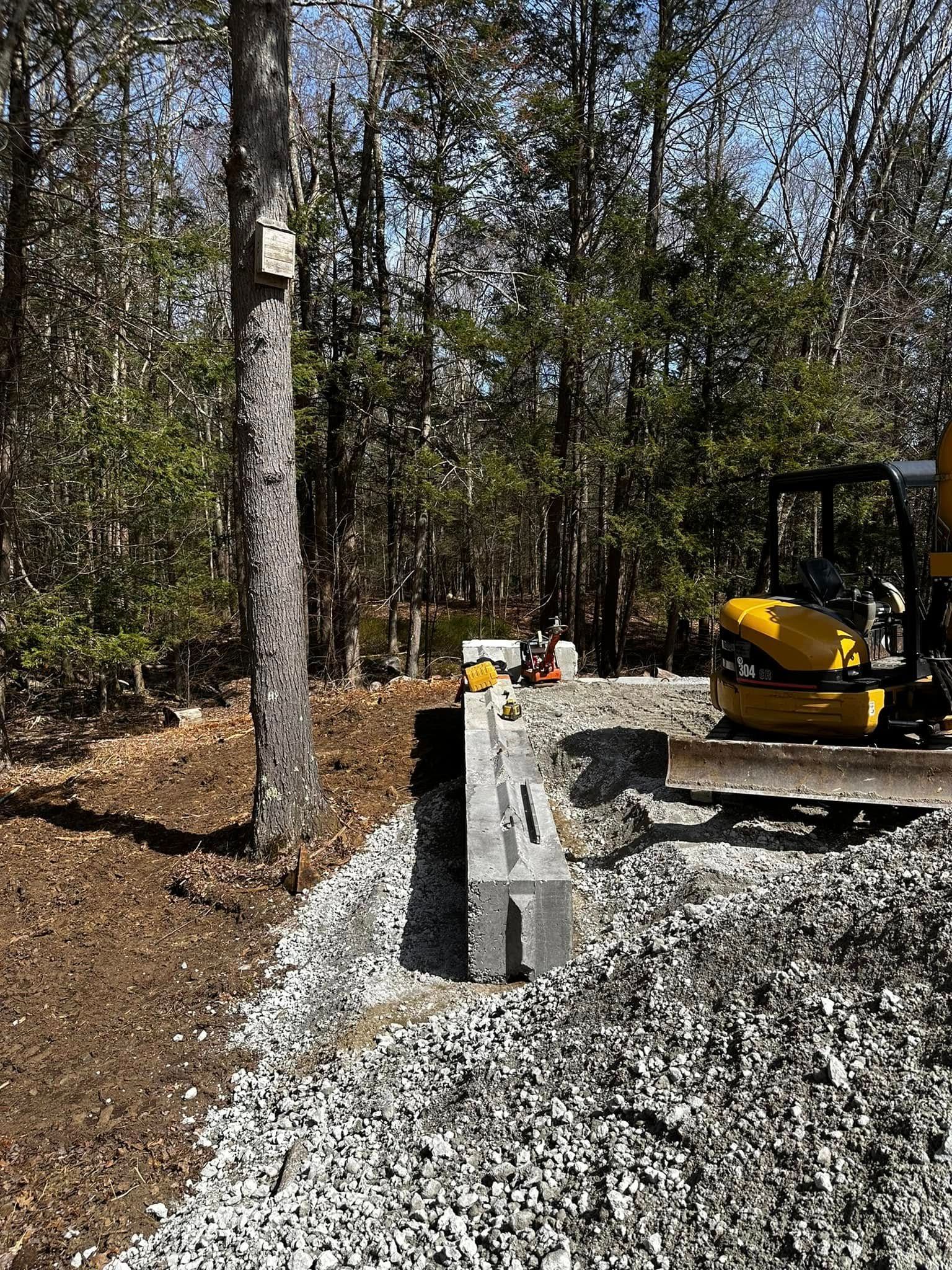 Construction site with retaining wall being built with backhoe in wooded area.