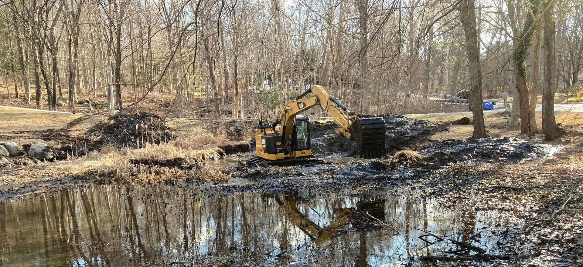 A yellow excavator working in a muddy pond in a wooded area. Trees in the background, reflections in the water.