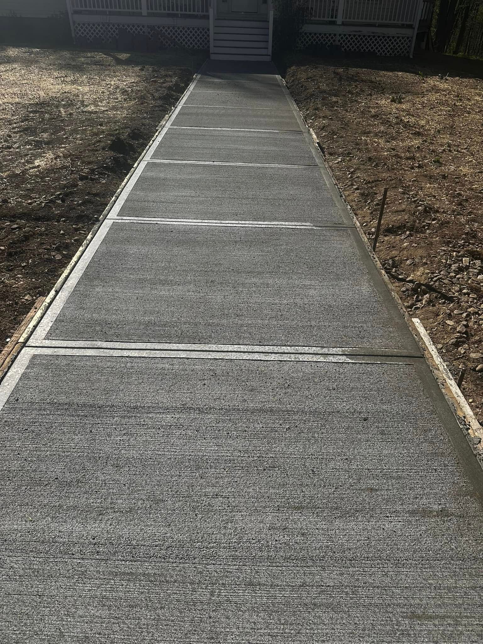 A newly poured concrete walkway with expansion joints leads toward a house with a porch.