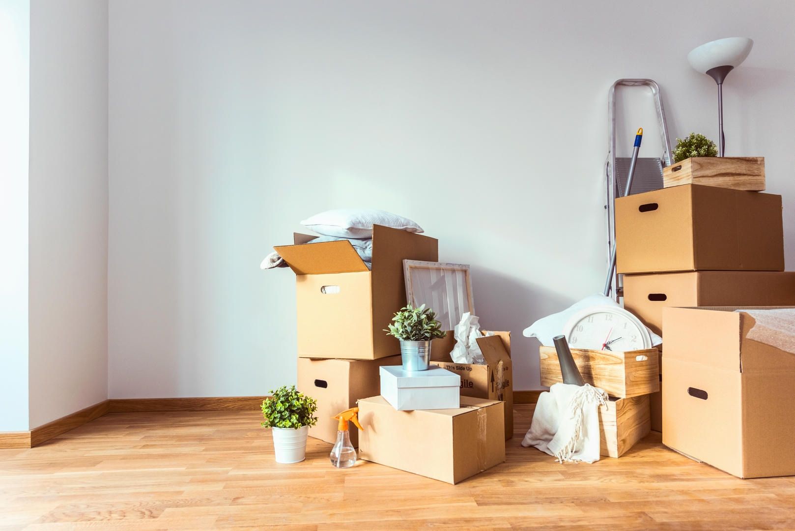 Cardboard boxes and belongings in a room with wood flooring. A ladder and a lamp are also in the room.