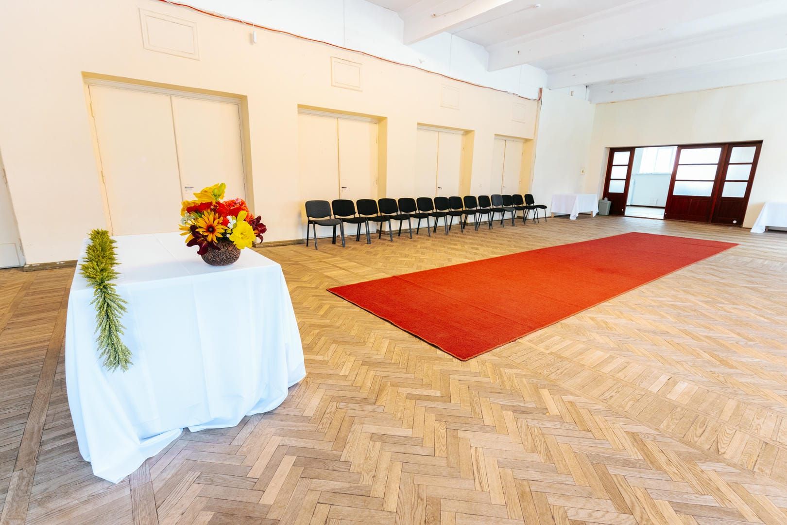 A white draped table with flowers sits in a room with a red carpet, chairs, and doors.