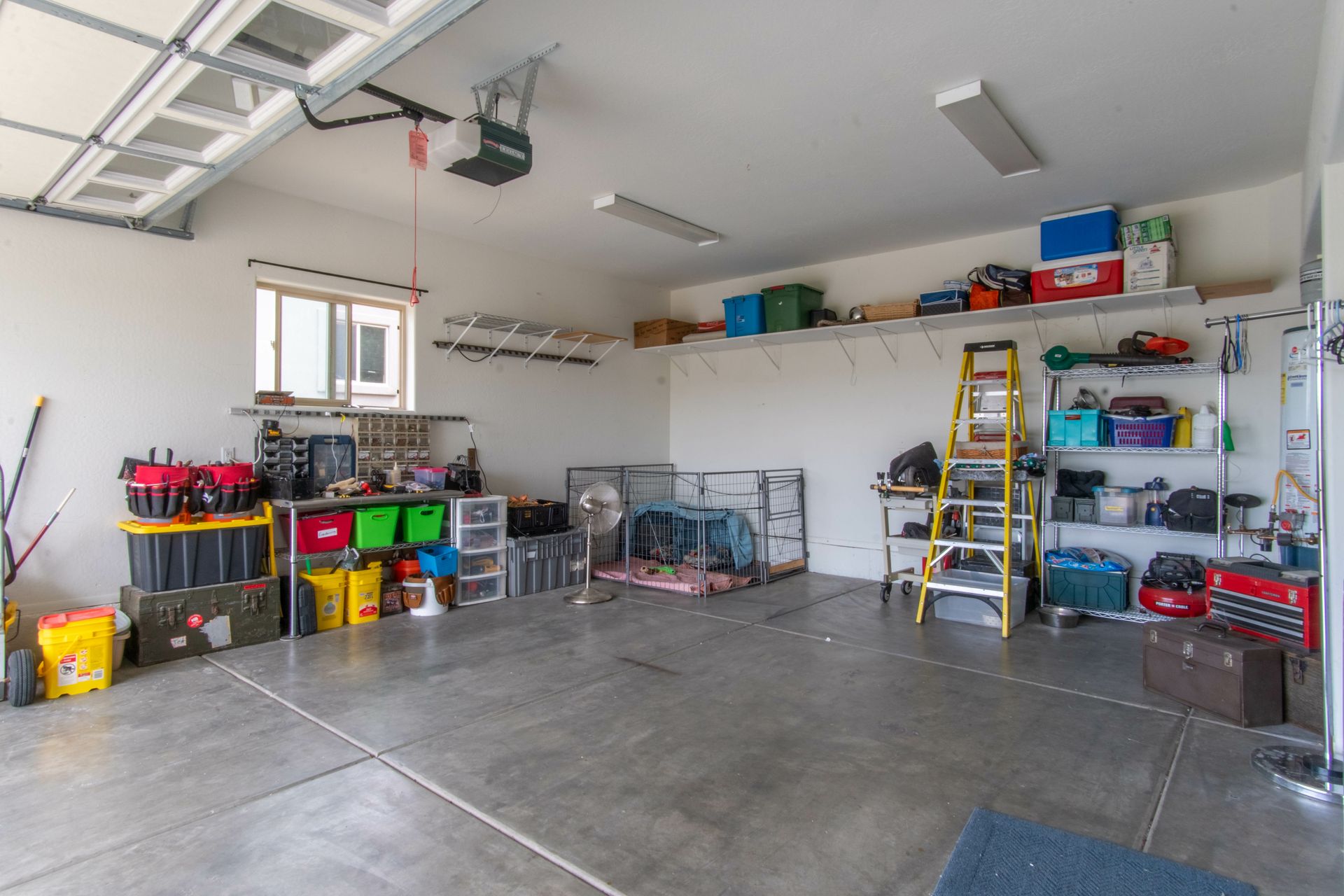 Cluttered garage with storage shelves and tools on concrete floor; white walls, garage door.