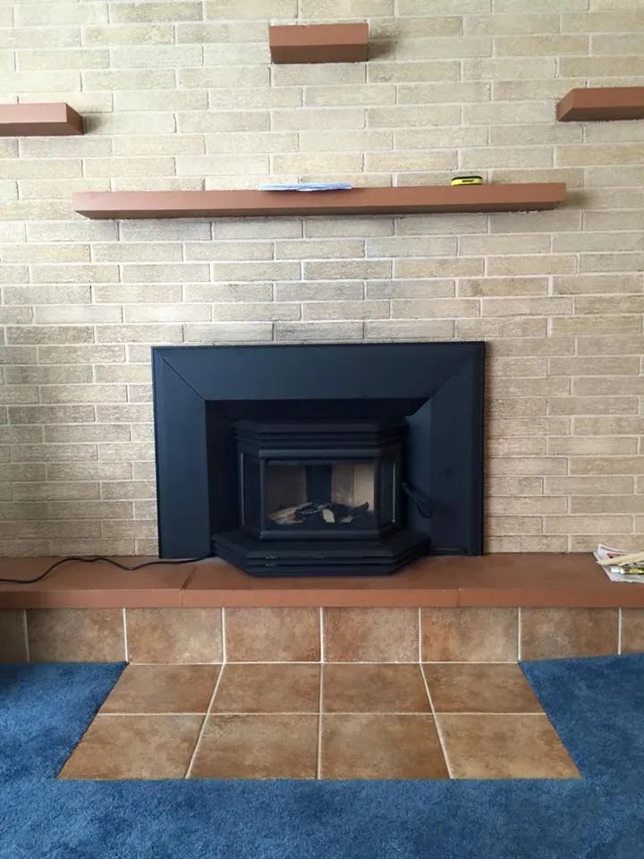 Living room fireplace with black surround, brick wall, wooden mantel shelves, and tiled hearth area