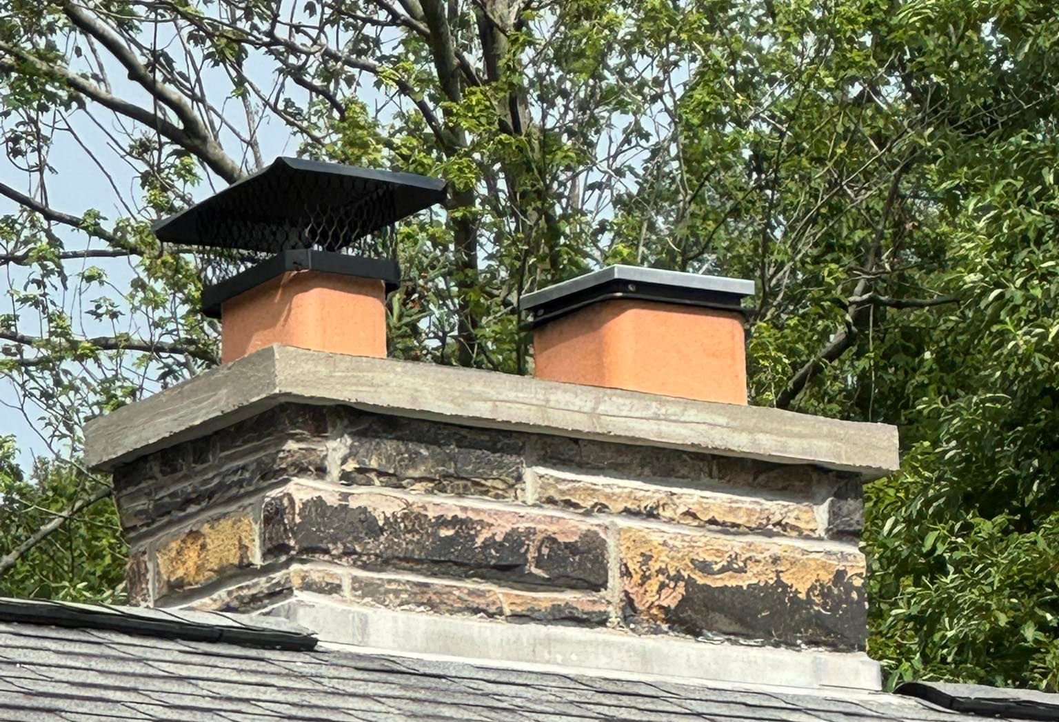 Brick chimney with two orange flues and black caps on a shingled roof, framed by green tree branches