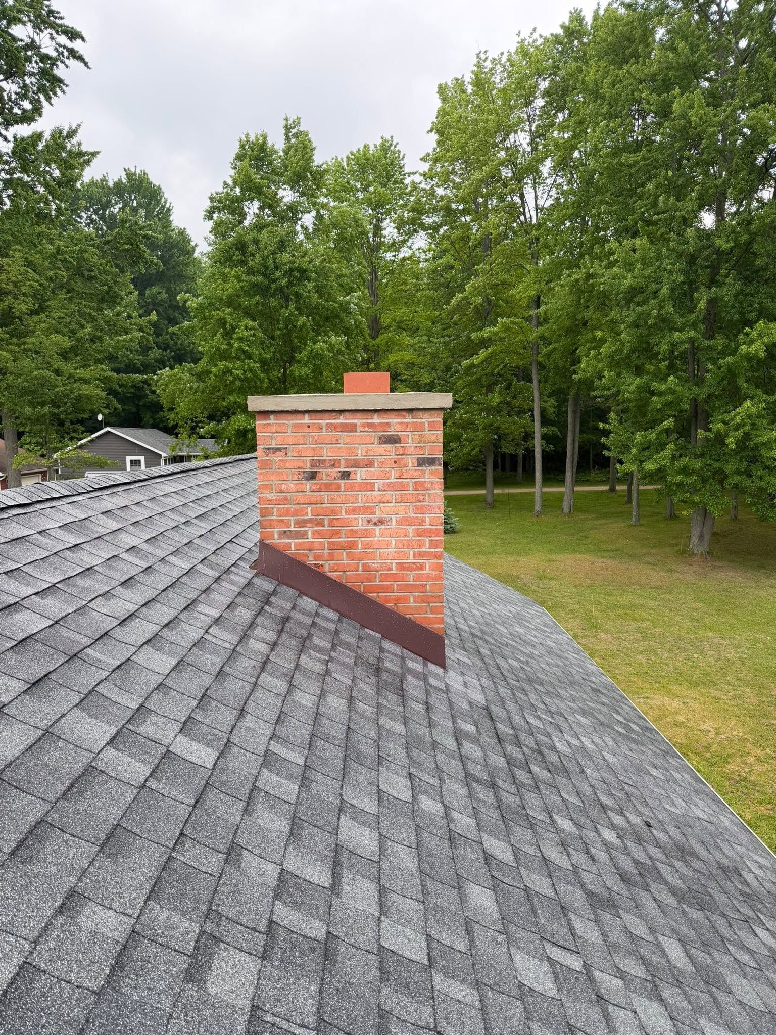 Gray shingled roof with a brick chimney, backed by green trees under an overcast sky