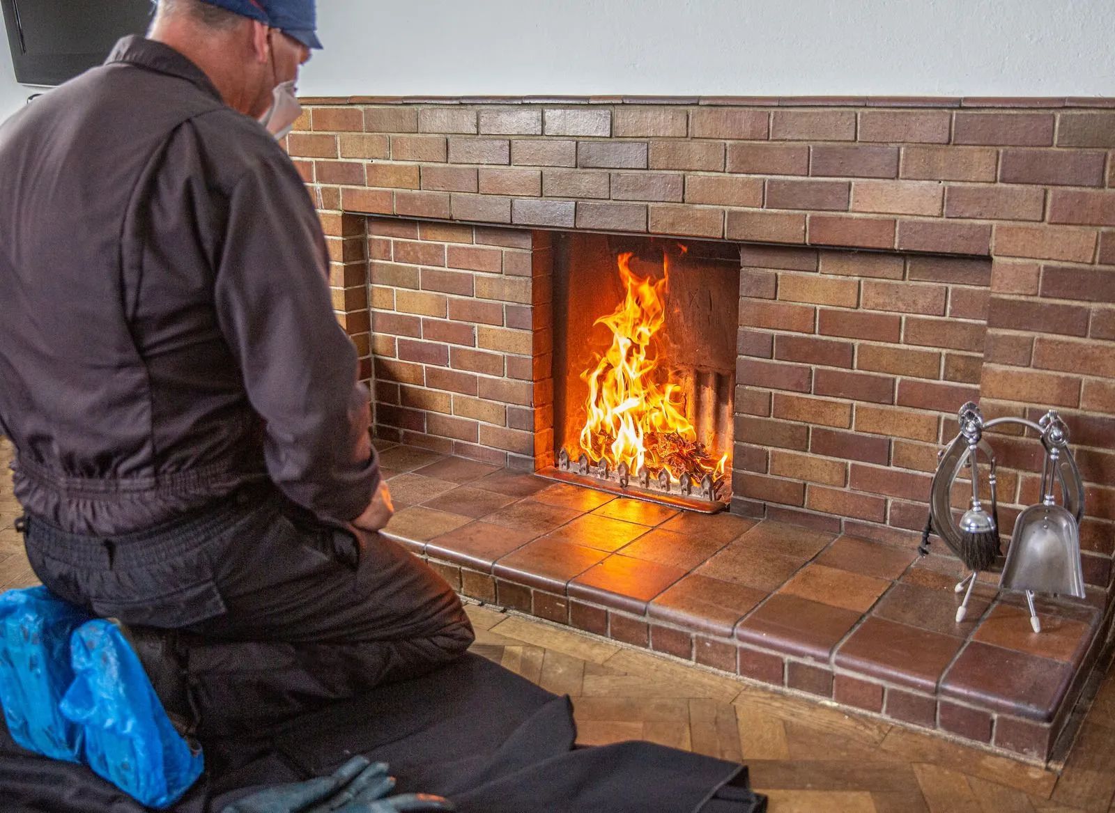 Person kneeling beside a lit brick fireplace with fire tools on the hearth