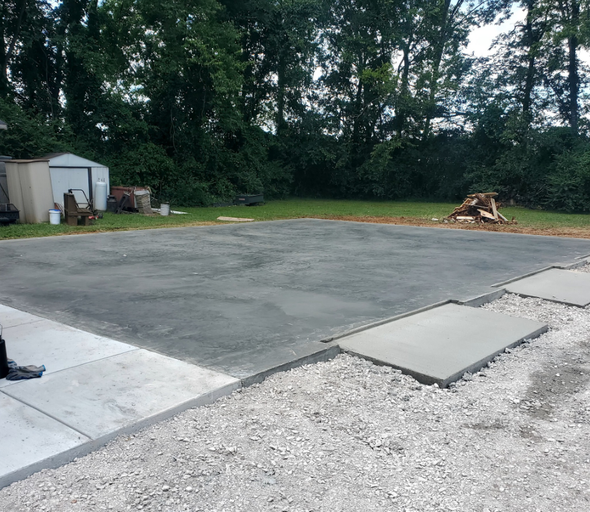 Newly poured concrete slab and sidewalk with gravel edging; shed and trees in background.