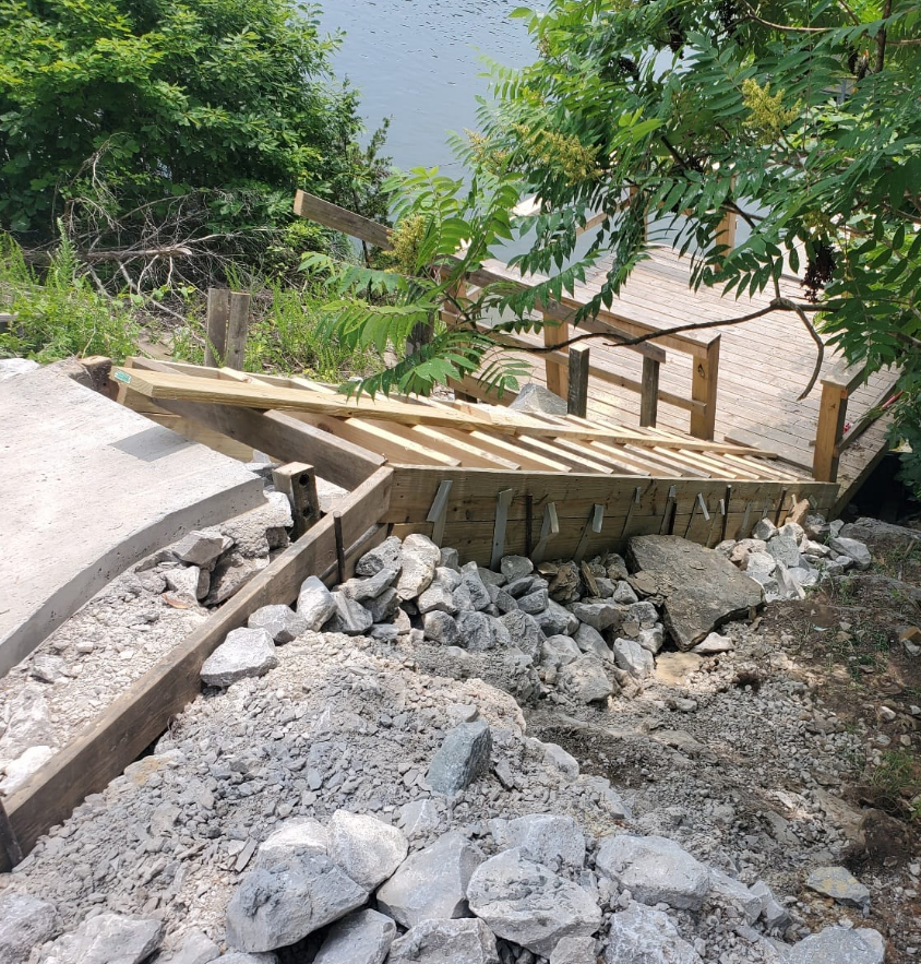 Construction site: stairs and ramp being built near water, surrounded by rocks and trees.
