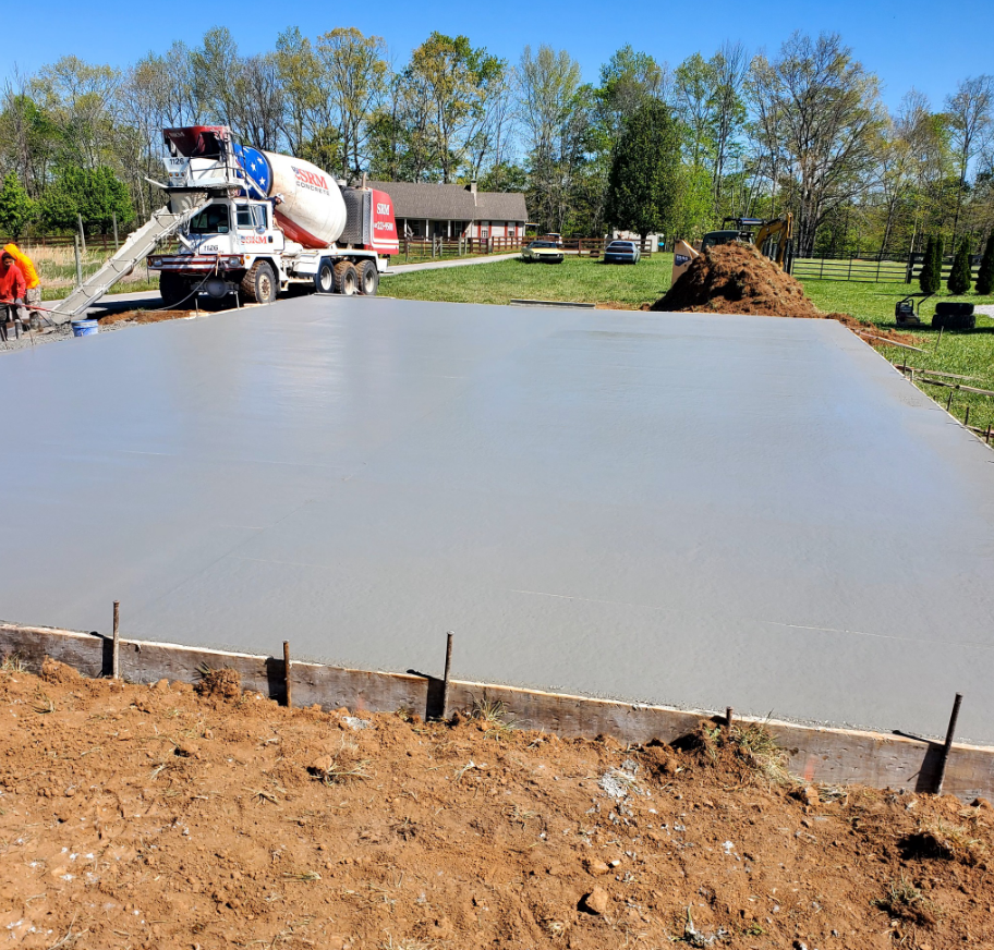 Concrete being poured for a foundation. Cement truck is in action. Brown ground, blue sky, and trees in background.