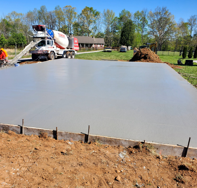 Concrete being poured from a truck, forming a smooth gray slab within wooden forms on a dirt lot.