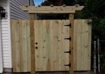 Wooden backyard gate and fence with a pergola-style top, attached to a house