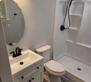 Compact white bathroom with sink, toilet, mirror, and walk-in shower with black fixtures